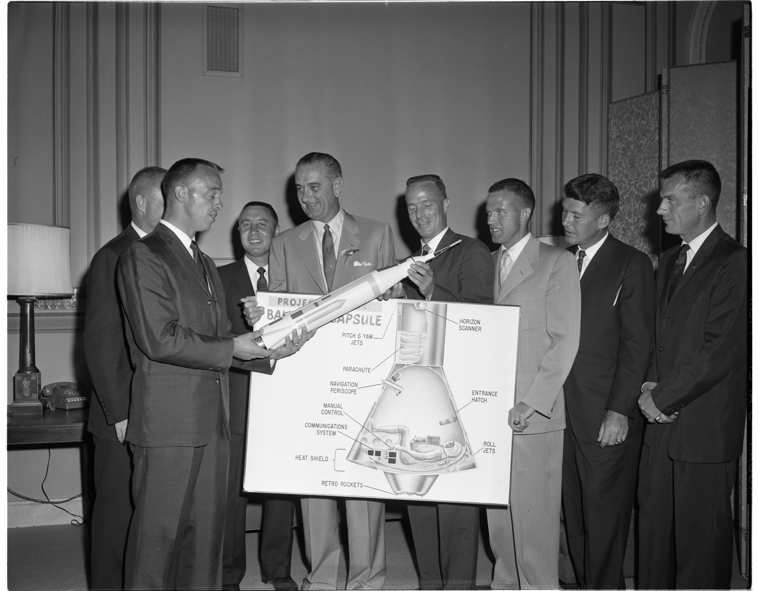 A group of men in suits hold a model rocket and a diagram of a space capsule. 